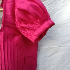 May include: A pink, pleated, sleeveless blouse with a gathered puff sleeve. The blouse has a round neckline and a fitted bodice.