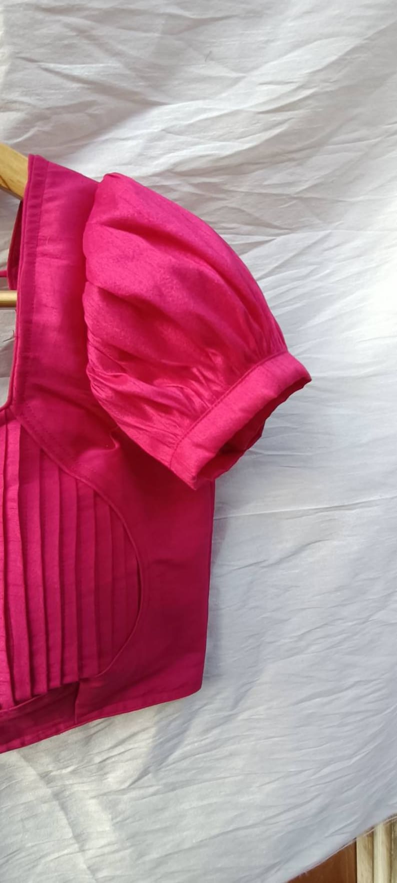 May include: A pink, pleated, sleeveless blouse with a gathered puff sleeve. The blouse has a round neckline and a fitted bodice.