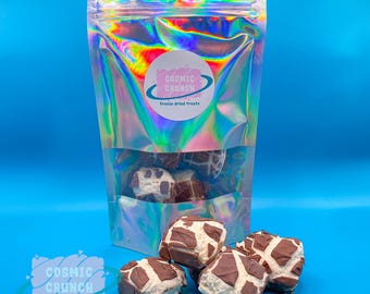 May include: A bag of freeze-dried treats from Cosmic Crunch. The bag is iridescent and has a pink and white logo with the text "Cosmic Crunch" and "Freeze dried treats". The bag is open and there are several freeze-dried chocolate squares on a blue surface.
