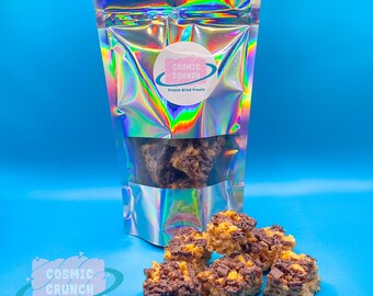 May include: A bag of freeze-dried treats called "Cosmic Crunch" with a holographic iridescent finish. The bag is open and there are several pieces of the treats scattered on a blue surface. The treats are a combination of chocolate and a light brown, crunchy cereal.