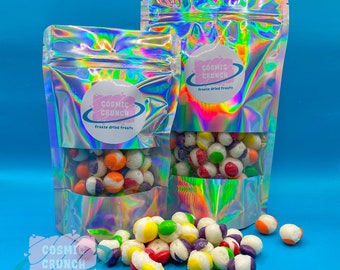 May include: Two iridescent bags filled with colourful freeze-dried fruit snacks. The bags have a white circle logo with the text "Cosmic Crunch" and "freeze dried treats". The snacks are scattered on a blue surface.
