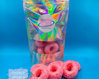 May include: A bag of pink freeze-dried donut treats with the logo "Cosmic Crunch Freeze Dried Treats" on the front of the bag.  There are also pink freeze-dried donuts on a blue surface.