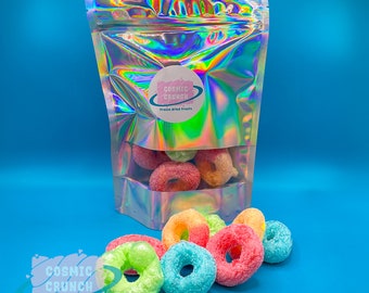 May include: A holographic bag filled with freeze-dried fruit snacks. The bag has a pink circle logo with the text "Cosmic Crunch" and "Freeze Dried Treats". The snacks are in the shape of colourful rings and are scattered on a blue surface.
