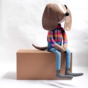 May include: A stuffed fabric dog toy with long brown ears, a white face, and a black nose. The dog wears a red, blue, and yellow plaid long-sleeve shirt, blue fabric pants, and grey lace-up boots. The toy has a brown tail and is sitting upright.