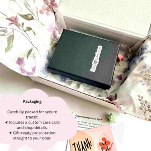 May include: A black rectangular jewelry box with a white handnpixel.com sticker, nestled inside a white gift box lined with floral tissue paper featuring pink and blue flowers, accompanied by a care card and a thank you note with floral art.