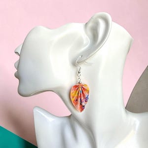 May include: A drop earring featuring a folded, petal-shaped charm made of multicolored paper with vibrant orange, yellow, pink, and blue patterns. The charm hangs from a silver-toned metal hook and a small silver bead, displayed on a white mannequin.