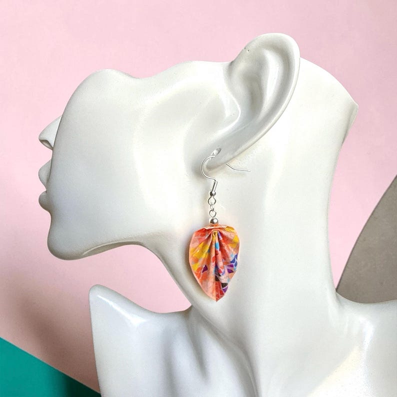 May include: A drop earring featuring a folded, petal-shaped charm made of multicolored paper with vibrant orange, yellow, pink, and blue patterns. The charm hangs from a silver-toned metal hook and a small silver bead, displayed on a white mannequin.