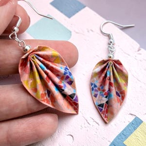 May include: A pair of drop earrings featuring folded leaf-shaped pendants made from paper with a colorful mosaic pattern of pink, orange, yellow, blue, and purple fragments. The earrings hang from silver-toned metal hooks with small silver beads.