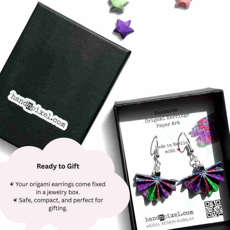 May include: A pair of handmade origami earrings featuring a fan-shaped design with a vibrant purple, green, and blue pattern. The earrings are displayed in a black jewelry box with a white card that reads Handmade Origami Earrings Paper Art.