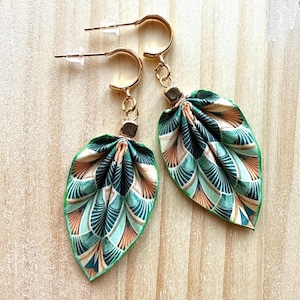 Peut inclure: Une paire de boucles d'oreilles créoles dorées avec des pendentifs en tissu en forme de feuille. Le tissu présente un motif géométrique en éventail dans des tons bleu sarcelle, vert foncé et pêche, avec une bordure verte. Chaque boucle a un fermoir.
