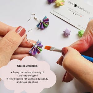 May include: A pair of fan-shaped origami earrings featuring multicolored paper patterns, suspended from silver-toned metal hooks. A paintbrush is applying a clear resin coating to the paper surface. A business card with the text handnpixel.com is visible.
