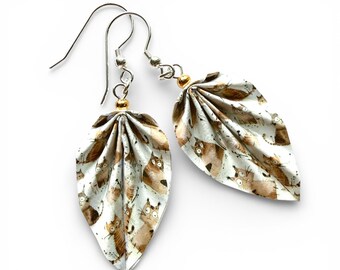 May include: A pair of dangle earrings featuring folded, leaf-shaped pendants made of off-white material with a repeating brown and black cartoon cat pattern. The pendants are attached to silver-toned metal hooks with small gold-toned beads.
