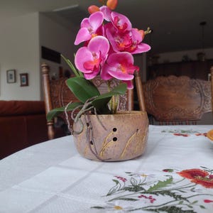 May include: A pink orchid plant in a brown ceramic pot with holes. The pot has a speckled glaze and is sitting on a white tablecloth with a floral pattern.