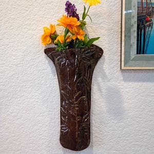 May include: A dark brown, glazed ceramic wall pocket vase with a flared top and embossed bamboo stalk patterns. The vase is filled with a small bouquet of bright orange marigolds and a single purple flower, creating a vibrant contrast against the dark finish.