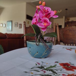 May include: A pink orchid plant in a blue ceramic pot with drainage holes. The plant is sitting on a white tablecloth with an embroidered floral design.