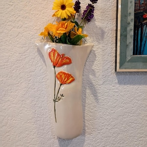 May include: A wall-mounted ceramic vase in a cream color, featuring two raised, hand-painted orange poppy flowers on the front. The vase is filled with a bouquet of yellow daisies and purple lavender stems, hanging against a textured white wall.