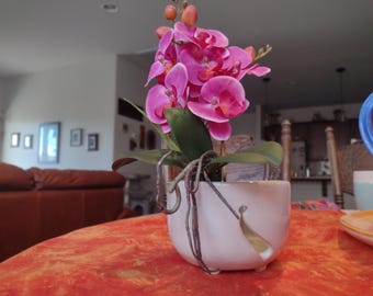 May include: A pink orchid plant in a white ceramic pot with a decorative metal accent. The plant is on a red and orange patterned table.