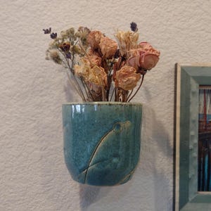 May include: A teal ceramic wall vase with a leaf design, holding dried roses and other flowers. The vase is mounted on a textured white wall, next to a framed picture of boats.