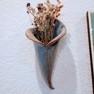 May include: A wall-mounted ceramic vase with a flared, horn-like shape and a glossy, speckled blue and brown glaze. The vase contains a bouquet of dried flowers in shades of beige, tan, and muted purple, extending upwards from the opening.