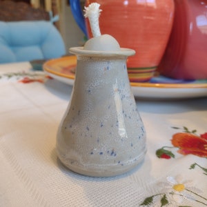 May include: A small, white ceramic oil lamp with a white wick and a speckled gray glaze. The lamp is sitting on a white tablecloth with floral embroidery.