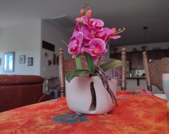 May include: A pink orchid plant in a white ceramic pot with a decorative cut-out design. The plant is sitting on a red and orange tablecloth.