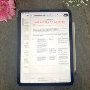 May include: A tablet displaying a document titled Characters of Macbeth with a table analyzing play characters. The screen shows a navigation sidebar, a top toolbar with an edit button, and a stylus attached to the side of the dark-colored device.