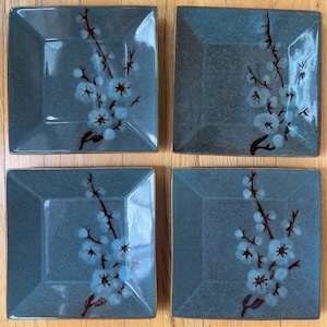 May include: Four square ceramic plates with a teal glaze. Each plate features a floral design with light blue blossoms and brown stems. The plates are arranged on a wooden surface, showcasing a cohesive set for dining or display.