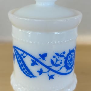 May include: White milk glass jar with a blue floral design and a white lid. The jar has a scalloped rim and a knob on the lid.
