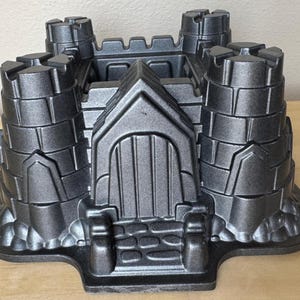 May include: A black metal castle-shaped cake pan with a detailed design. The pan has four towers, a central structure with a door, and a base with a stone-like texture.