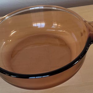 May include: A clear, amber-colored Vision cookware bowl with two handles. The bowl has a rounded shape and a black rim. The Vision logo and other text are visible on one of the handles. This durable bowl is suitable for cooking and serving.