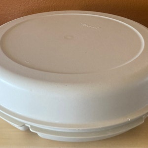 May include: A light beige, round Tupperware container with a lid. The lid has a slightly recessed center and the word "Tupperware" is printed on the top. The container has a ridged edge.
