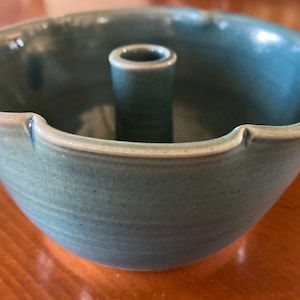 Puede incluir: Un cuenco de cerámica de color turquesa con un soporte cilíndrico central. El cuenco tiene un borde festoneado y un acabado liso y esmaltado. La pieza es probablemente hecha a mano y adecuada para uso decorativo o funcional.