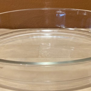May include: A clear, oval-shaped glass baking dish with two handles. The dish has a slightly raised rim and is designed for oven use. The handles have a decorative, ridged design. The dish is empty and sits on a light-colored surface.