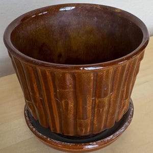May include: A brown ceramic planter with a bamboo-style design. The planter has a wide opening and sits on a matching saucer. The glossy finish reflects light, highlighting the textured vertical lines. A decorative piece for indoor or outdoor use.