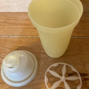 May include: A yellow plastic shaker cup with a white lid and a white plastic insert with six holes. The shaker cup is designed for mixing protein shakes or other beverages.