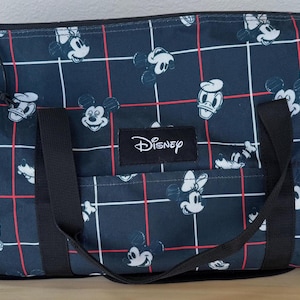 May include: A navy blue duffel bag featuring a repeating pattern of white Mickey Mouse and Minnie Mouse faces, along with Donald Duck. The bag has a red and white grid design, black handles, and a Disney logo.