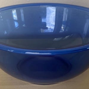 May include: A deep blue ceramic mixing bowl with a rounded shape and a wide rim. The bowl is empty and reflects the surrounding environment. The bowl is sitting on a light-colored wooden surface.