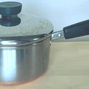 May include: Stainless steel saucepan with a lid and a black handle. The pot has a copper-colored bottom and a black knob on the lid. The handle has a loop for hanging. The pot is sitting on a light-colored wooden surface.