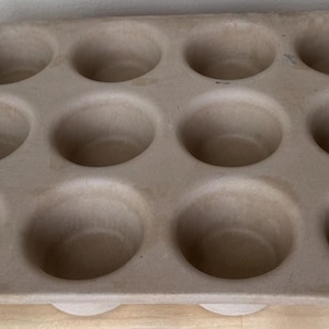 May include: A tan-colored, rectangular muffin pan with twelve round cavities. The pan is made of a heavy material and sits on a light-colored wooden surface. The cavities are evenly spaced and appear to be about 3 inches in diameter.
