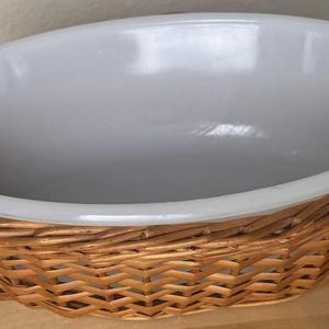 May include: Oval-shaped serving dish with a light gray interior and a woven wicker basket base. The dish has a smooth, glossy interior and a wide rim. The wicker base is made of light brown woven material.
