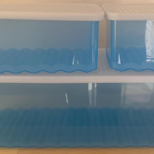 May include: Three blue plastic storage containers with white lids are stacked on a wooden surface. The containers have a wavy design along the top edge. The containers are designed for organizing items.