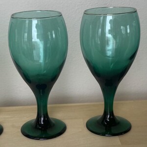 May include: Four emerald green wine glasses. The glasses have a classic design with a rounded bowl and a long stem. The base and stem are a darker shade of green than the bowl. The glasses are arranged in a row on a light-colored surface.
