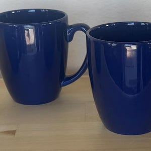 May include: Four navy blue ceramic mugs with curved handles are arranged on a light brown wooden surface. The mugs have a glossy finish and a simple, modern design, suitable for coffee or tea.