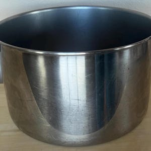 May include: A stainless steel cooking pot with a wide opening and two black handles. The pot has a cylindrical shape and a shiny, reflective surface. The interior is dark, and the pot appears to be empty. The pot is sitting on a light-colored wooden surface.