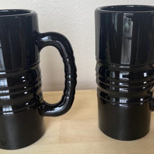 May include: Two black ceramic mugs with a glossy finish. Each mug has a cylindrical body with horizontal ridged detailing and a large, curved handle. The mugs are displayed on a light-colored wooden surface.