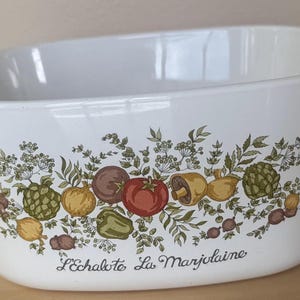 May include: White casserole dish with a decorative border of colorful vegetables and herbs. The design includes tomatoes, onions, mushrooms, and artichokes. The text "L'Echalote La Marjolaine" is printed on the dish.