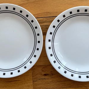 May include: Two white ceramic plates with a black square and line design around the rim. The plates are round and sit on a wooden surface. The plates are empty.