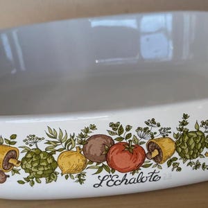 May include: White oval baking dish with handles, decorated with a colorful border of vegetables and herbs. The design includes tomatoes, mushrooms, and artichokes. The text "L'échalote" is printed on the dish.