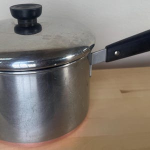 May include: A stainless steel saucepan with a lid and a black handle. The lid has a black knob. The handle has a metal ring at the end. The saucepan is sitting on a wooden surface.