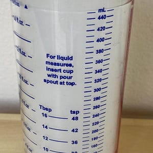 May include: Clear plastic measuring cup with a white lid. The cup has markings for fluid ounces, tablespoons, and teaspoons, as well as milliliters. Text on the cup reads "For liquid measures, insert cup with pour spout at top."
